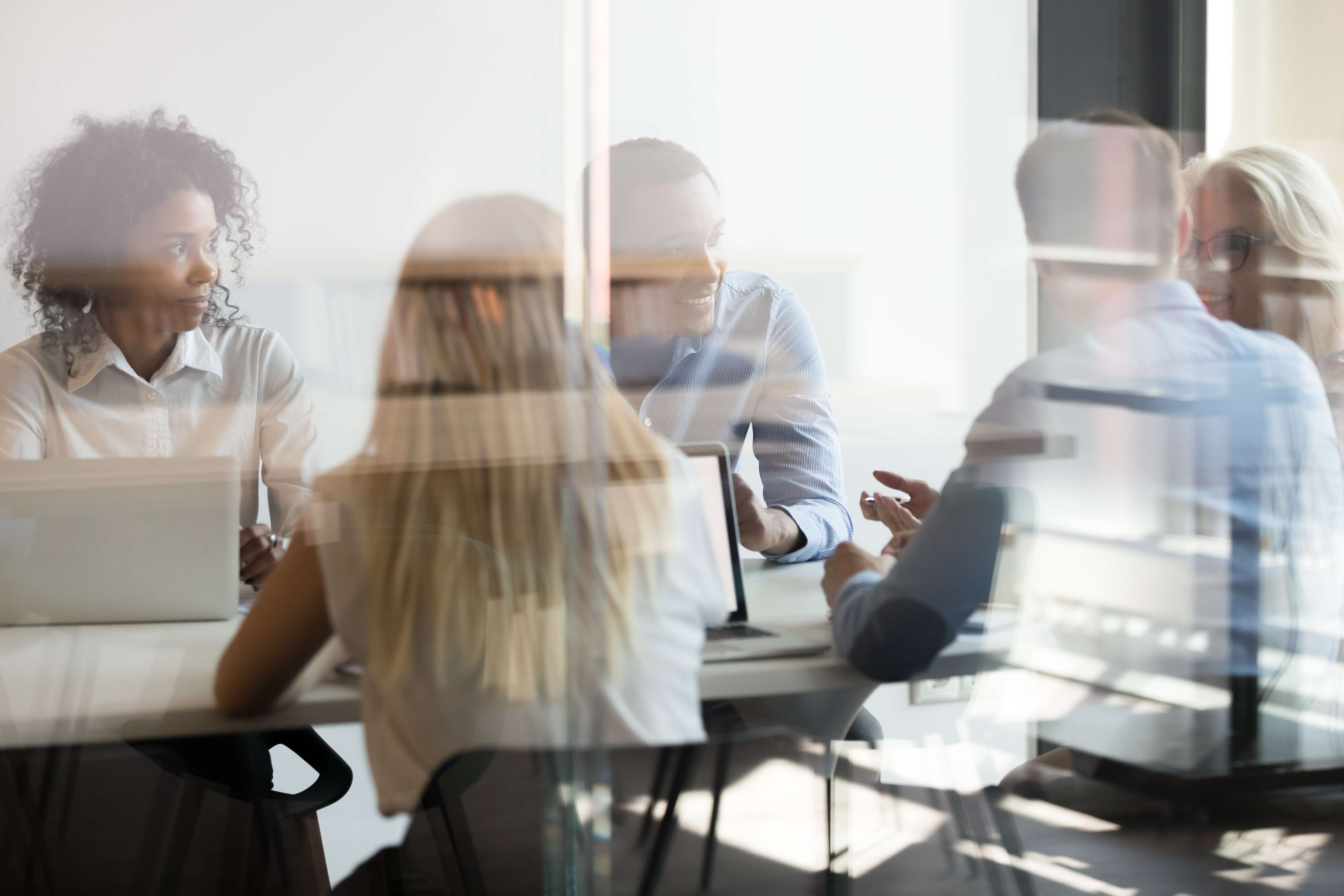 Four people are sitting around a table behind a glass partition in an office environment