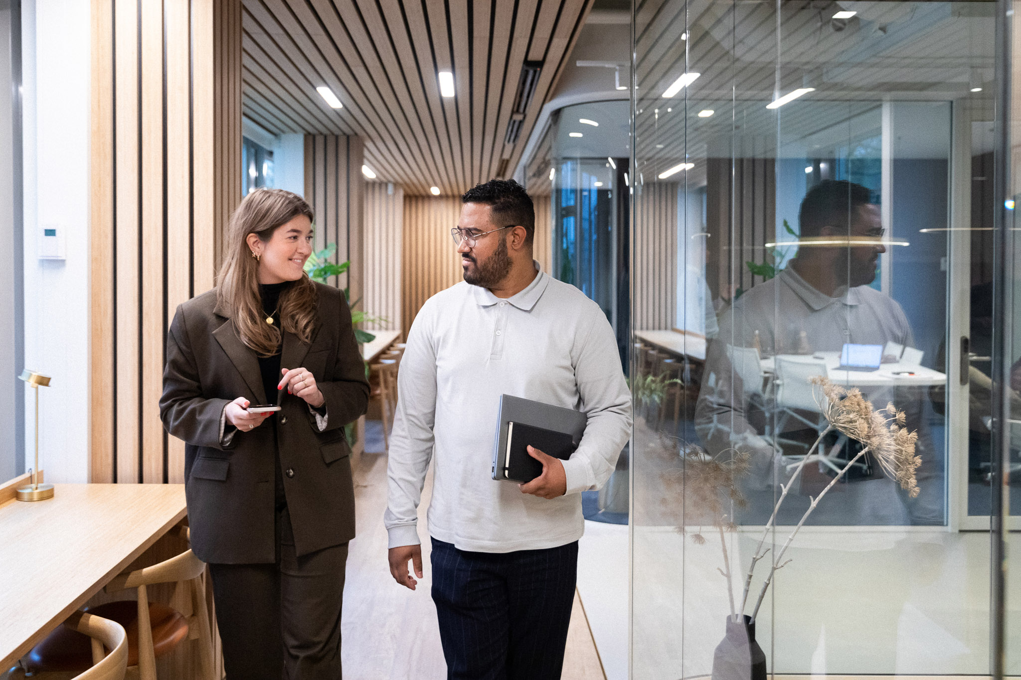 Two colleagues walking and talking in a modern office corridor. A woman in a dark blazer holds a phone while a man in a light grey top carries a laptop or tablet. They appear relaxed and engaged in conversation. The background features glass walls, wooden panels, and bright office interiors.