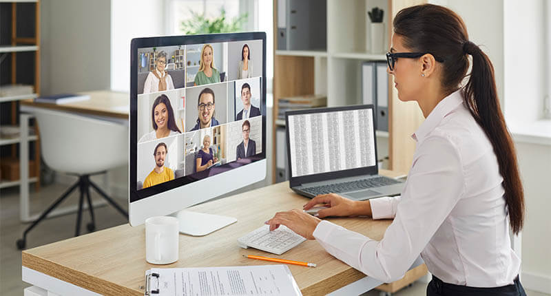 Female web conferencing administrator sitting at office table with desktop computer during virtual team meeting of company workers. Online business communication, telework and remote teamwork concepts
